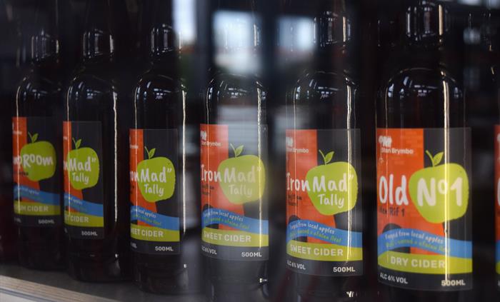Rows of dark bottles in a fridge with labels on. The labels are for different types of cider. They have a picture of an old blast furnace in orange an